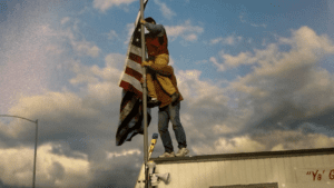 A photo of two men on the roof of a trailer house mounting an American flag. One man is on the shoulders of the other man.