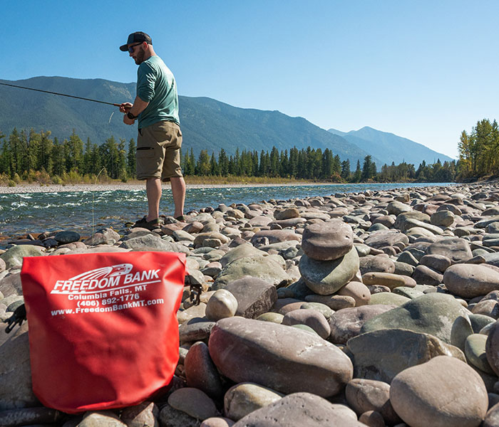 Bright red drybag with Freedom Bank logo in foreground. Man flyfishing at river with mountains in background.